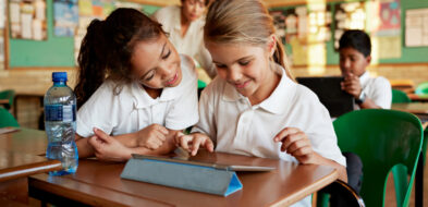 Two smiling elementary age girls using a tablet while sharing a school desk